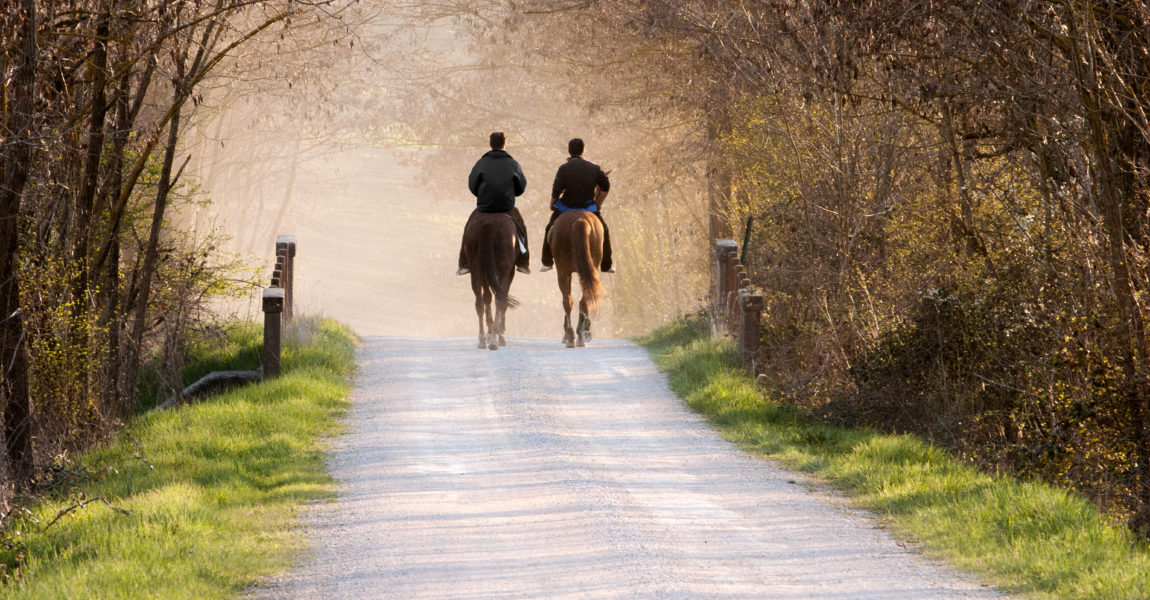 Two man riding horses in dirt road at sunset Two man riding horses through a bridge in a dirt road of the countryside of tuscany with a beautiful warm lightening, Siena, Italy 