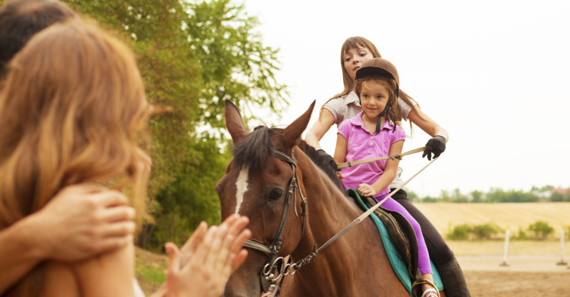 Child Riding Horse Outdoors. Little girl with riding hat learning how to ride a horse. Riding horse together with her instructor. Mother and father watching and encourage her. 