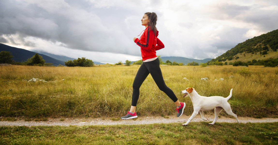 Jogging in the beautiful nature Young woman running in sports clothing with her pet dog. She is wearing a red sweater, doing exercise, in the beautiful nature, under the cloudy sky. Healthy lifestyle concept, keeping body in shape. Slightly toned image in analog / film colour. 
